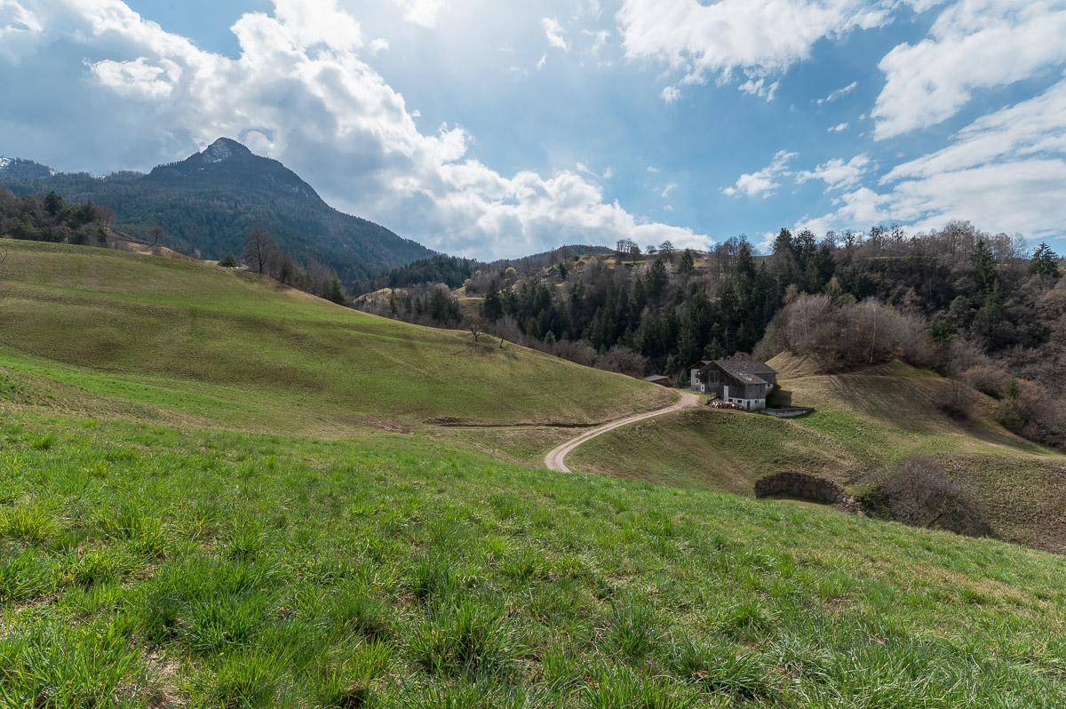 Grüne Wiesen erstrecken sich bis zu den Gebäuden am Hang, mit majestätischen Bergen im Hintergrund unter blauem Himmel.