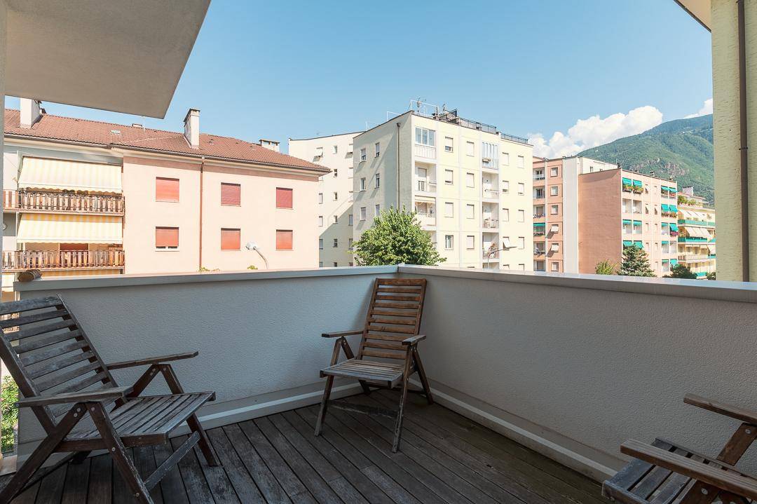 Sonniger Balkon mit Holzboden und Holzstühlen, bietet Blick auf die umliegenden Gebäude.