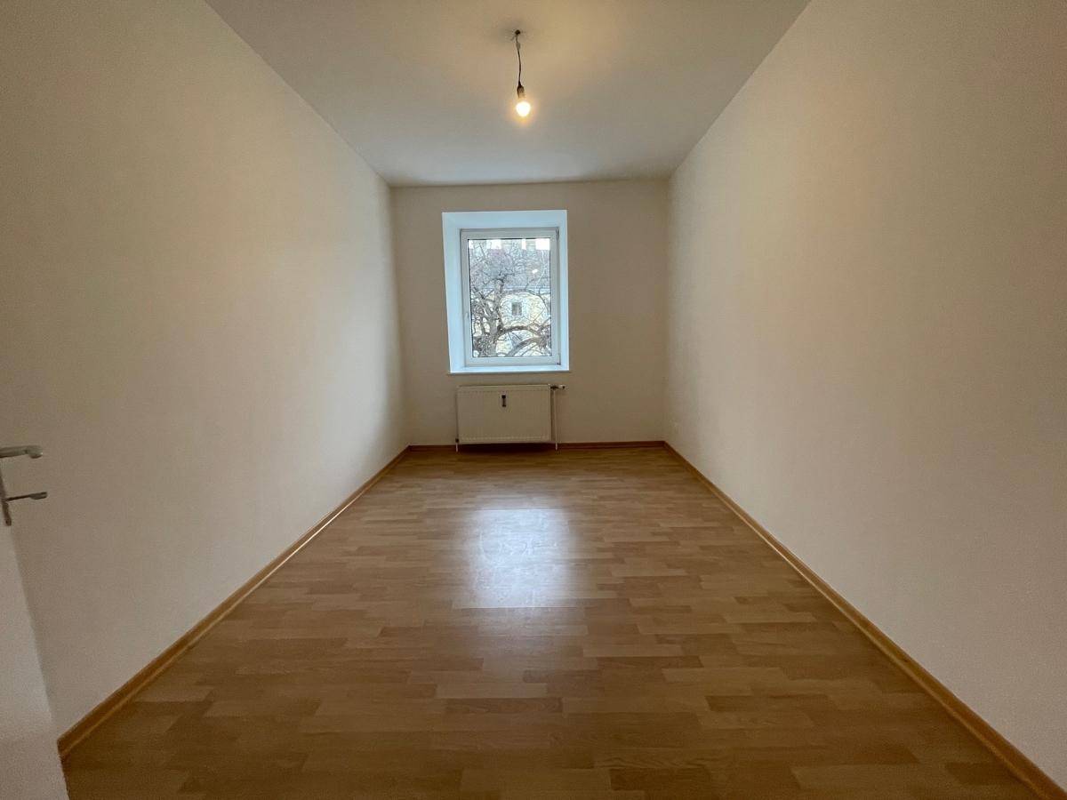 Langes, schmales Zimmer mit Parkettboden und einem Fenster mit Blick nach draußen.
