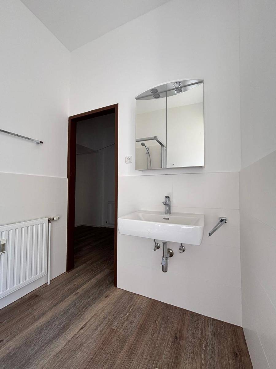 Badezimmer mit Waschbecken, Spiegelschrank und Heizkörper, das einen Zugang zu einem weiteren Raum bietet.