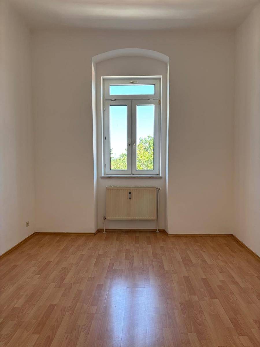 Leeres Zimmer mit Laminatboden und einem zentralen, hohen Fenster, das den Raum erhellt.