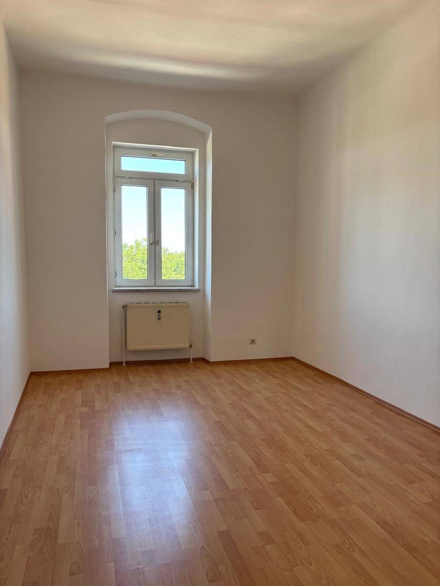 Unmöbliertes Zimmer mit Laminatboden und einem hohen Fenster, das den Raum mit Tageslicht füllt.