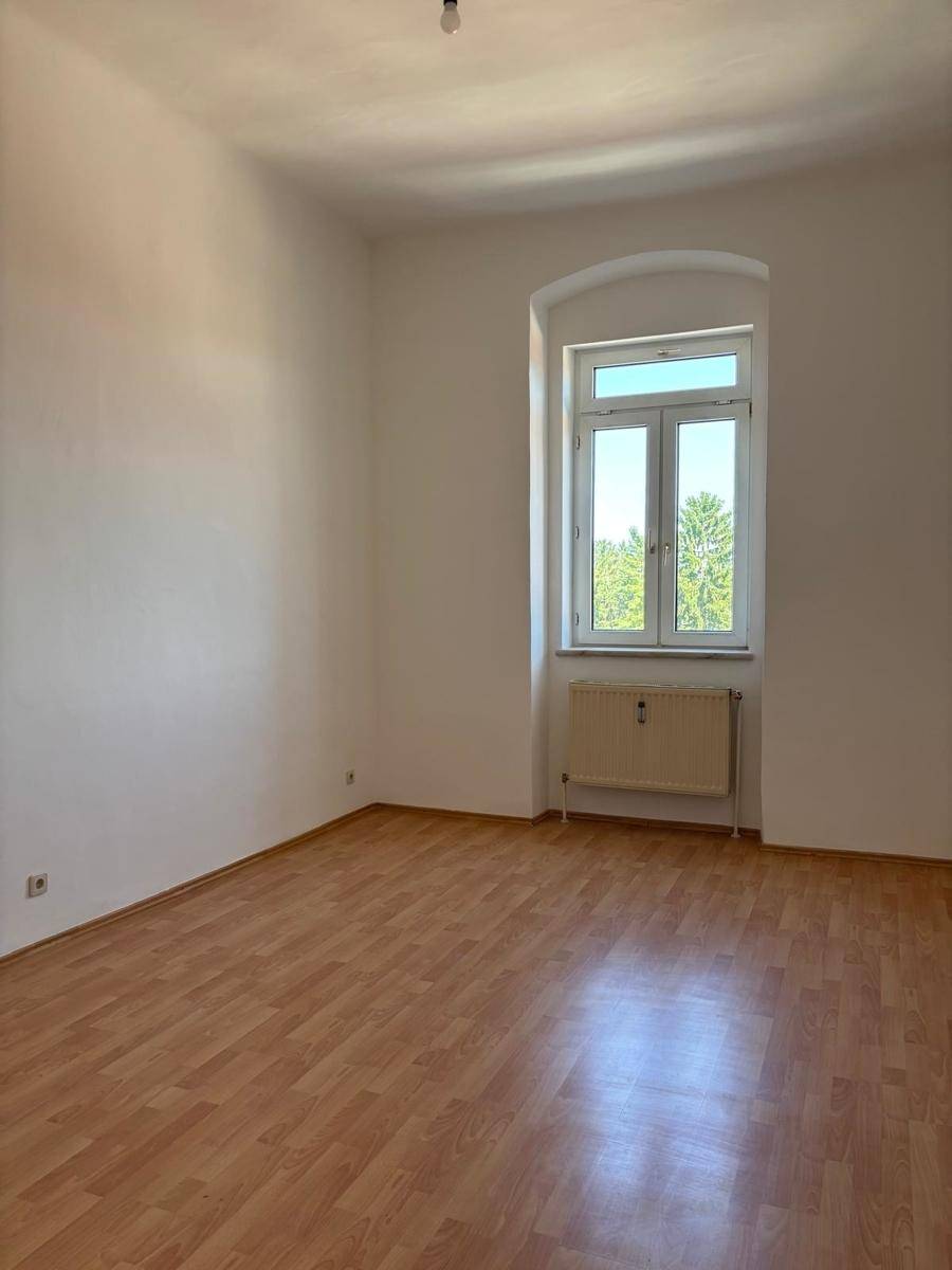Helles, leeres Zimmer mit warmem Laminatboden und einem hohen Fenster, das viel Licht spendet.