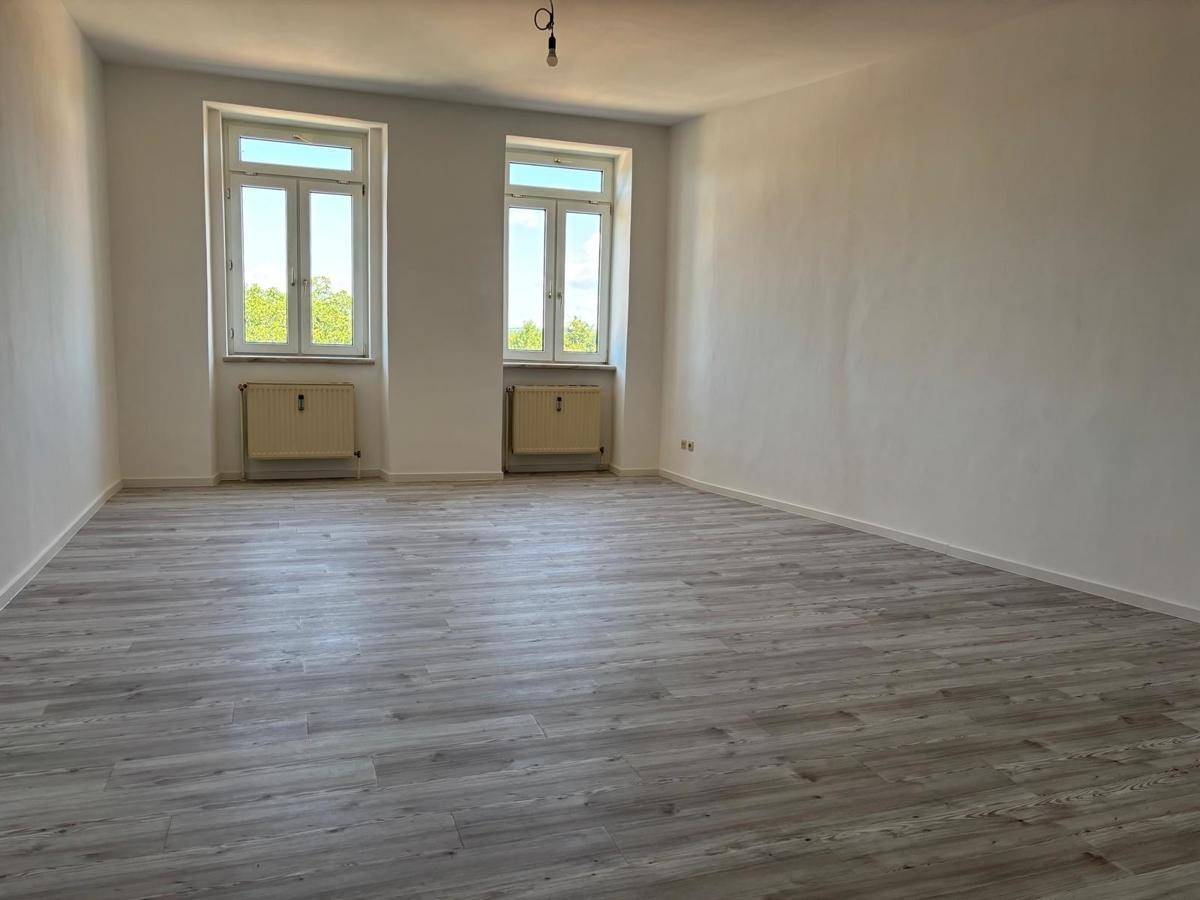 Geräumiges, unmöbliertes Zimmer mit hellem Laminatboden und zwei großen Fenstern für natürliche Beleuchtung.