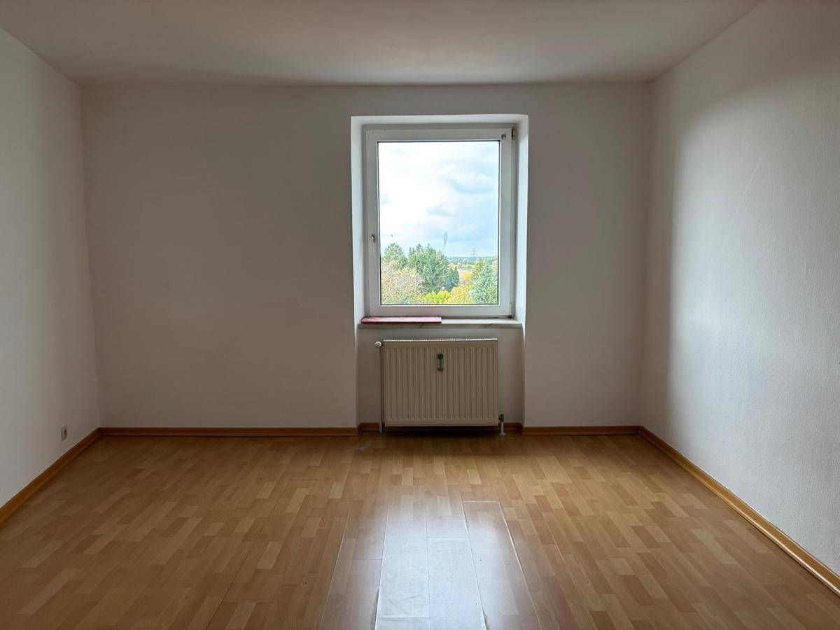 Lichtdurchflutetes Zimmer mit Laminatboden, weißen Wänden und einem Fenster mit Ausblick ins Grüne.