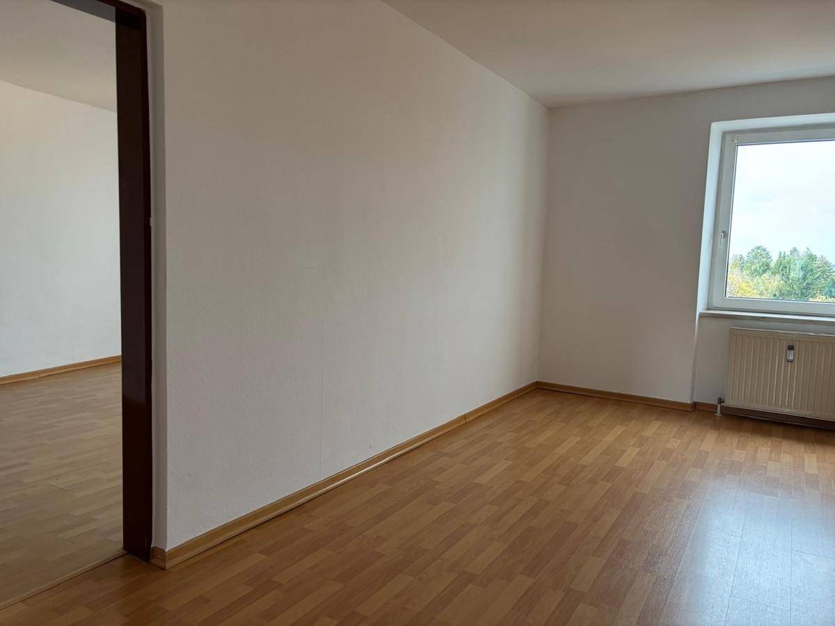 Geräumiges Zimmer mit hellem Laminatboden und Fenster, das eine grüne Aussicht bietet.