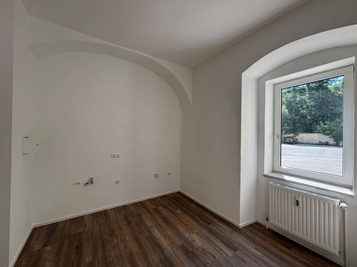 Leerer Raum mit Laminatboden und einem großen Bogenfenster, das viel Tageslicht hereinlässt.