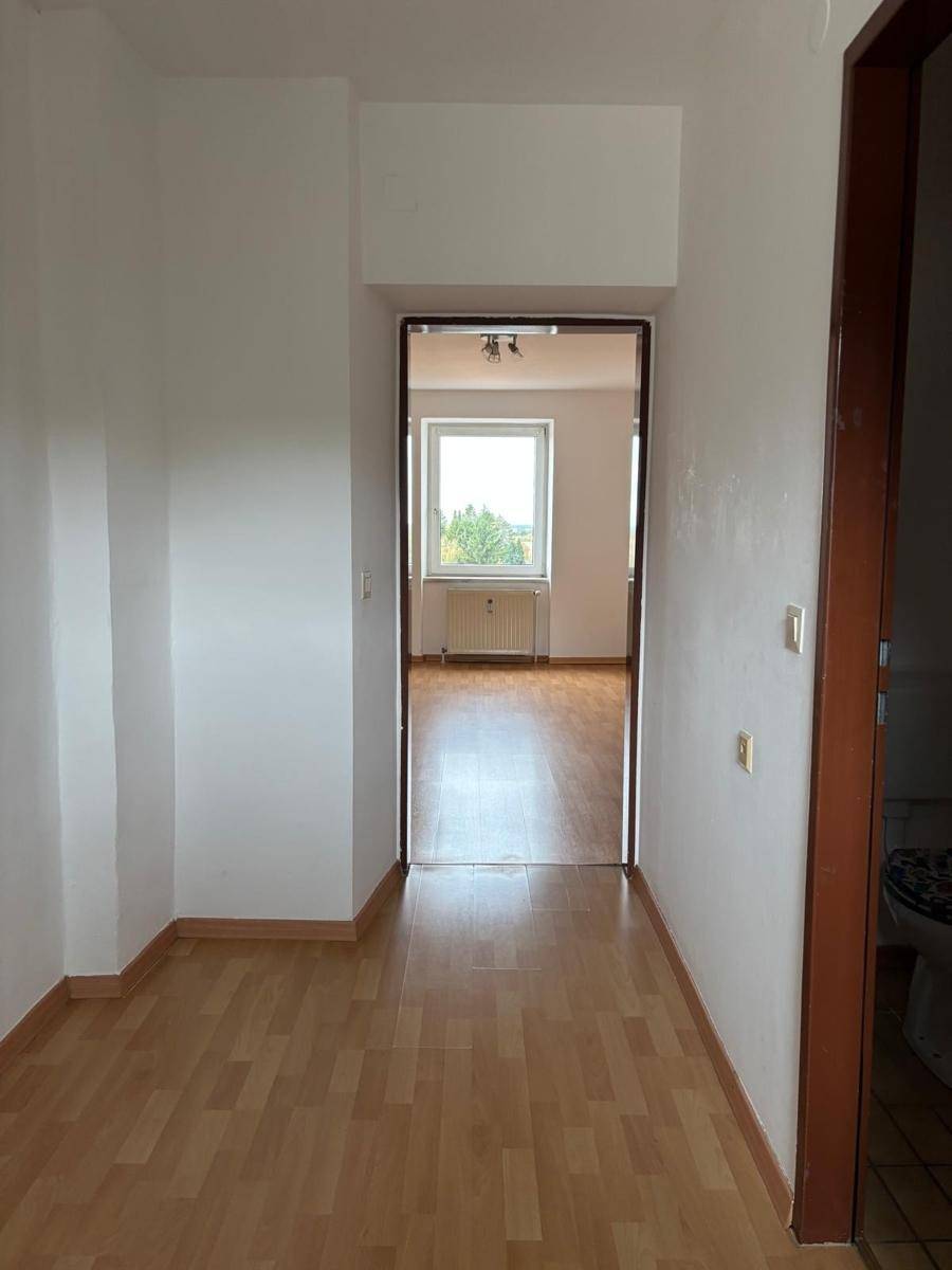 Flur mit Laminatboden, der zu einem hellen Zimmer mit Fenster und grüner Aussicht führt.