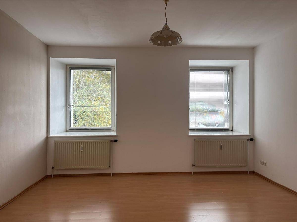 Helles, leeres Zimmer mit zwei Fenstern, Heizkörpern und Laminatboden, bereit zur individuellen Gestaltung.