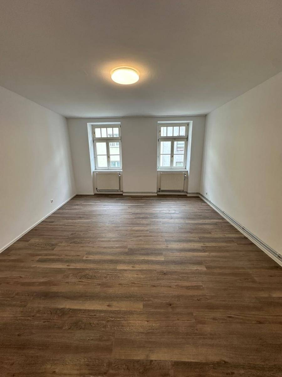 Leerer Raum mit Laminatboden, weißen Wänden und zwei Fenstern, die viel Licht spenden.