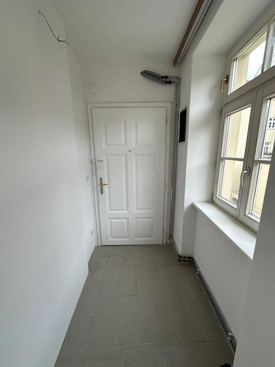 Heller Flur mit weißer Tür, Fliesenboden und einem Fenster, das Tageslicht hereinlässt.