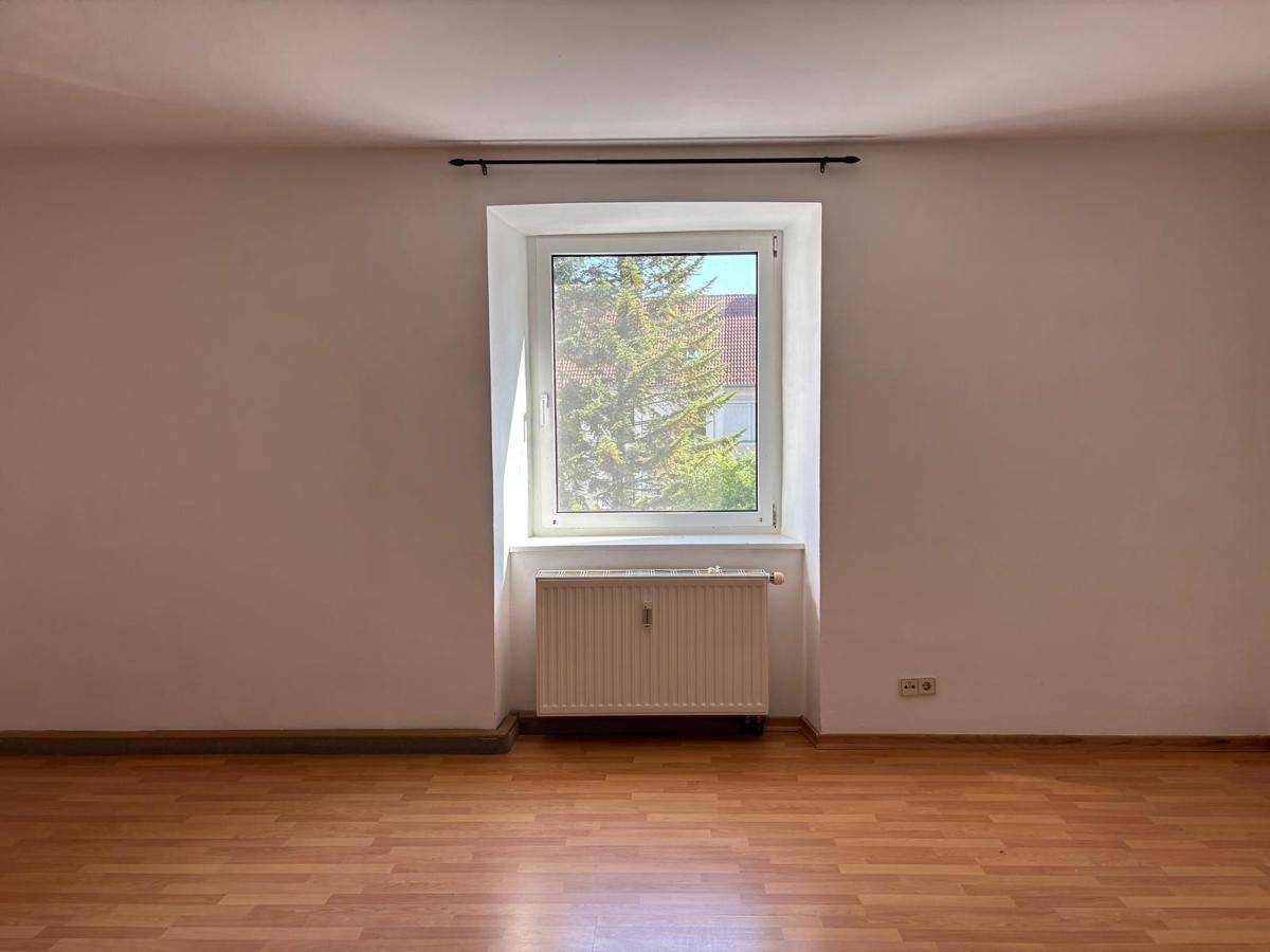 Leeres Zimmer mit Laminatboden, Fenster und Heizkörper, bietet viel natürliches Licht.