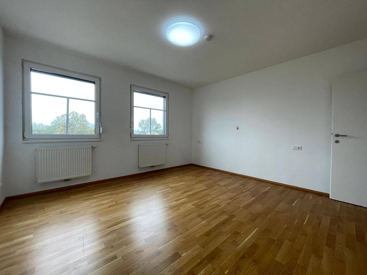 Leeres Zimmer mit Parkettboden, zwei Fenstern und einer Deckenleuchte, bietet viel Tageslicht.