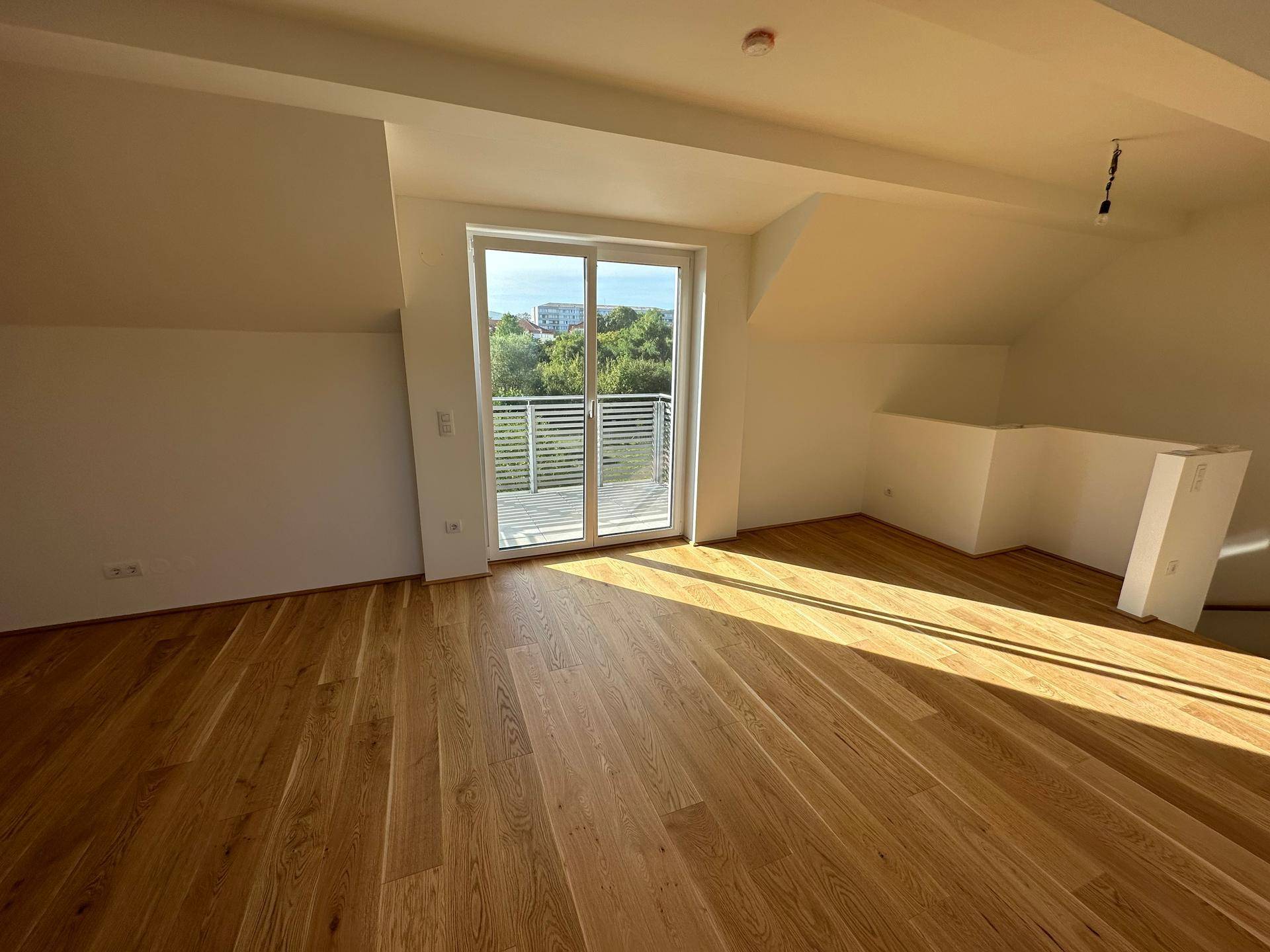 Heller Wohnraum im Dachgeschoss mit Holzboden, großen Fenstern und Zugang zum Balkon.