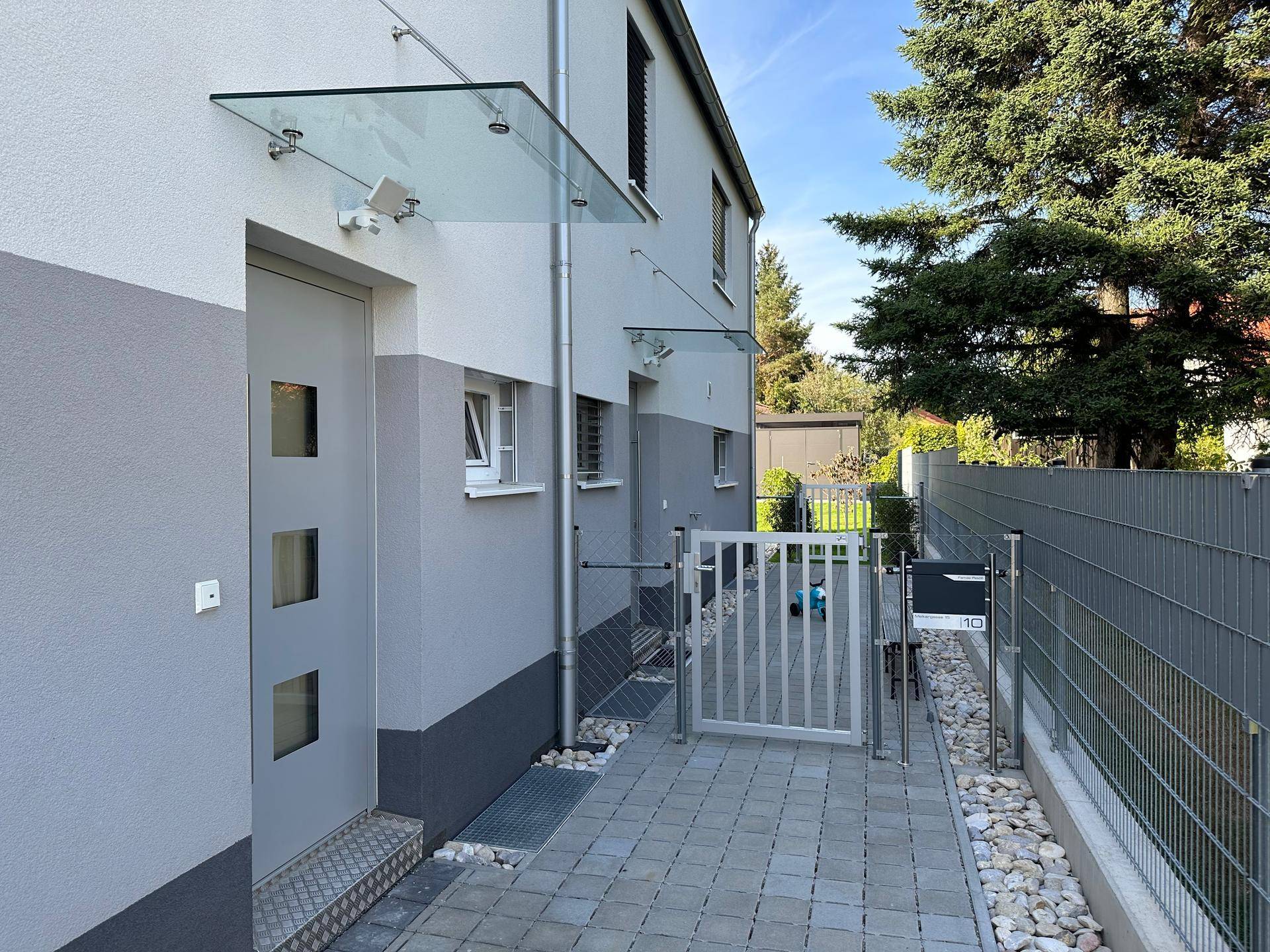 Eingangsbereich eines modernen Reihenhauses mit Glasvordach, gepflastertem Weg und Zaun.