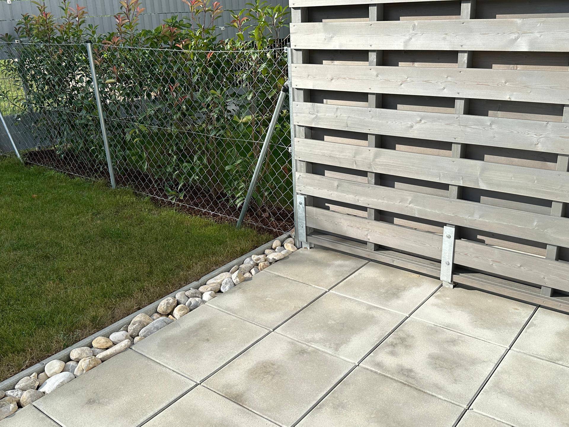 Ecke einer gepflasterten Terrasse mit Ziersteinen, Rasen und einem modernen Holzzaun.