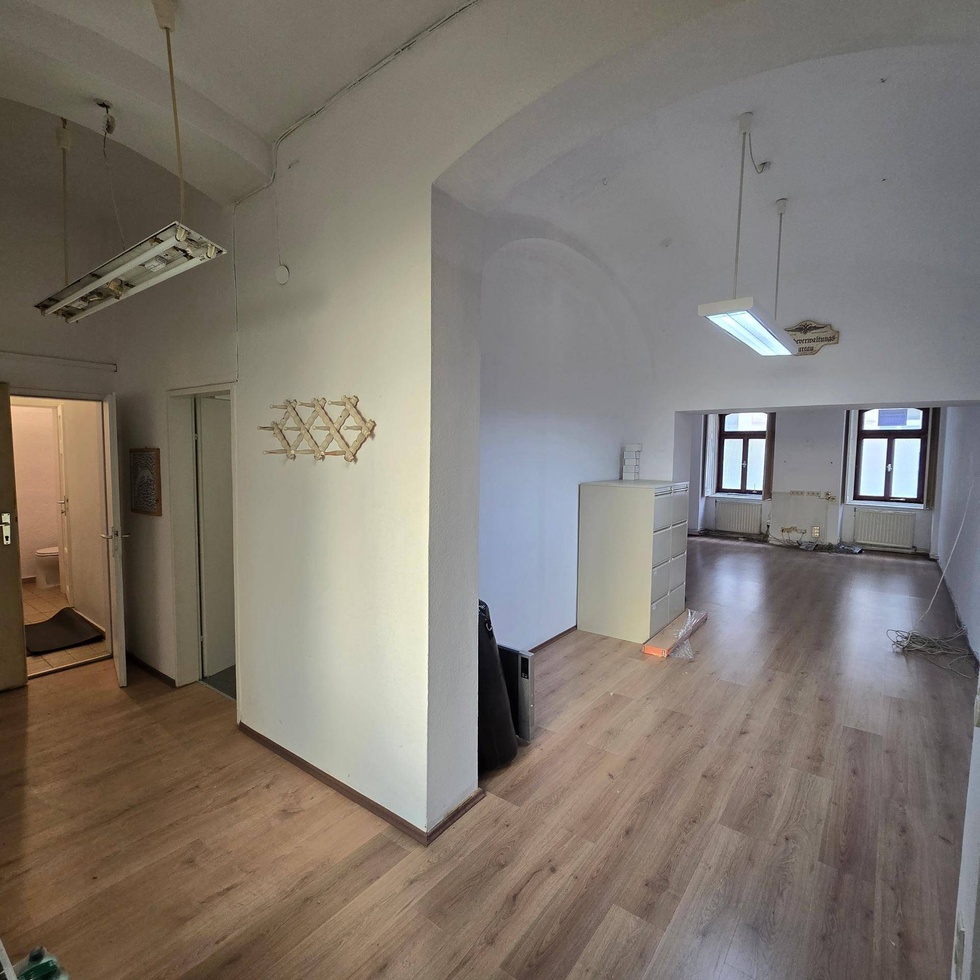 Großer Büroraum mit Laminatboden, gewölbter Decke und Blick in weitere Räume, ausgestattet mit Deckenleuchten.
