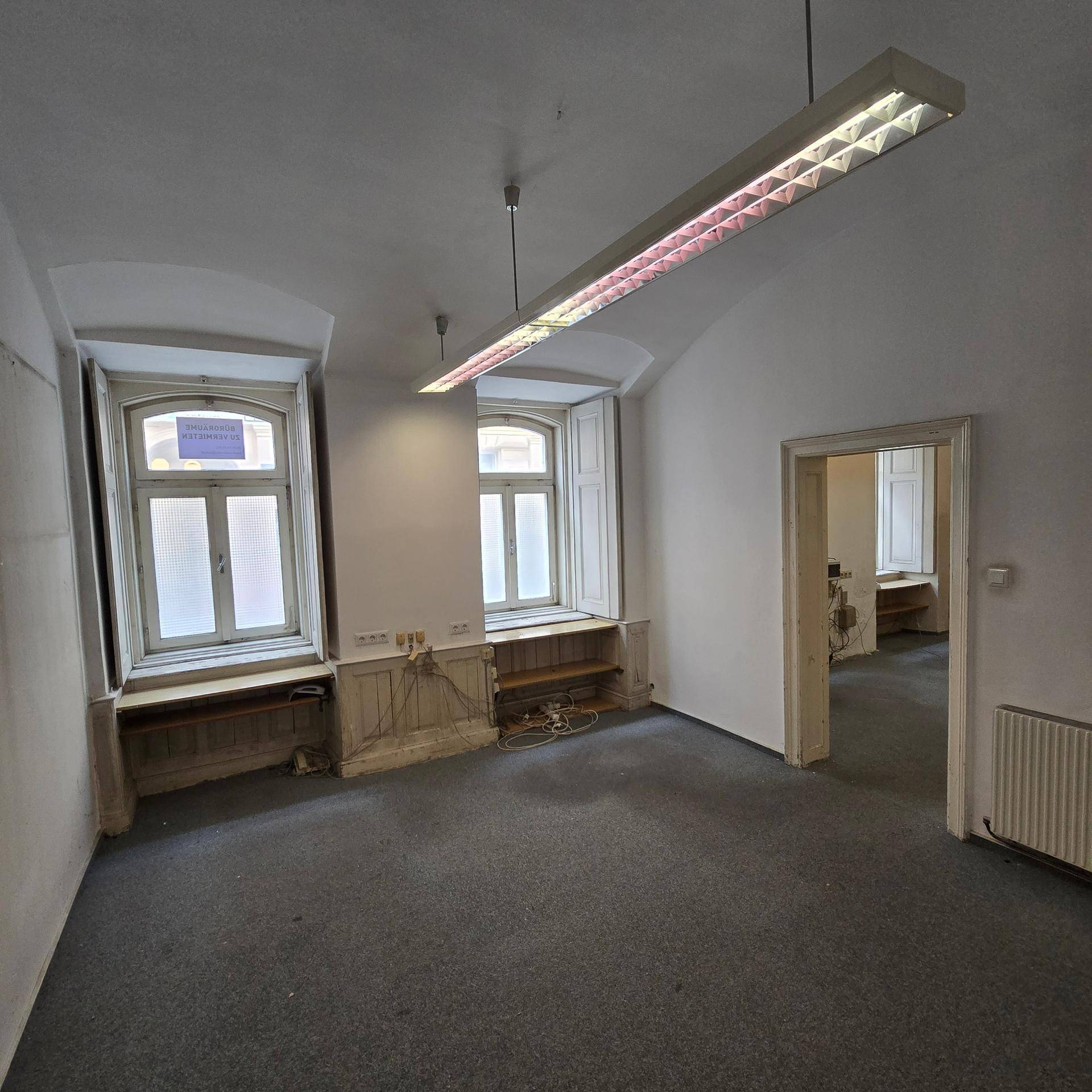 Büroraum mit Teppichboden, zwei Fenstern mit Jalousien und einer Deckenleuchte, in gutem Zustand.