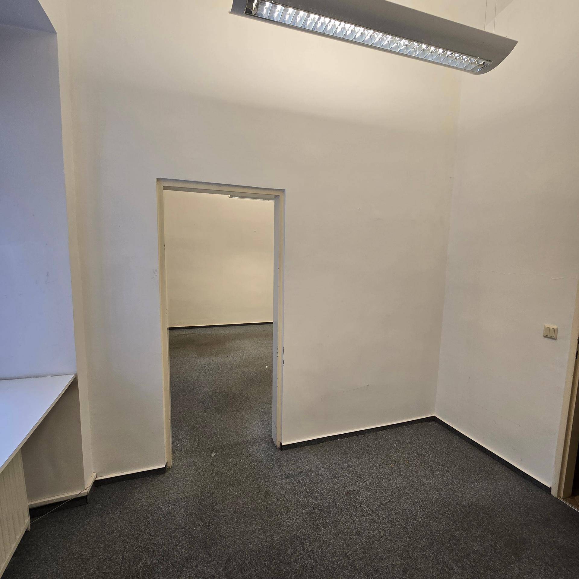 Kleiner Büroraum mit Teppichboden und einer offenen Tür zu einem weiteren Bereich, beleuchtet von einer Leuchtstofflampe.