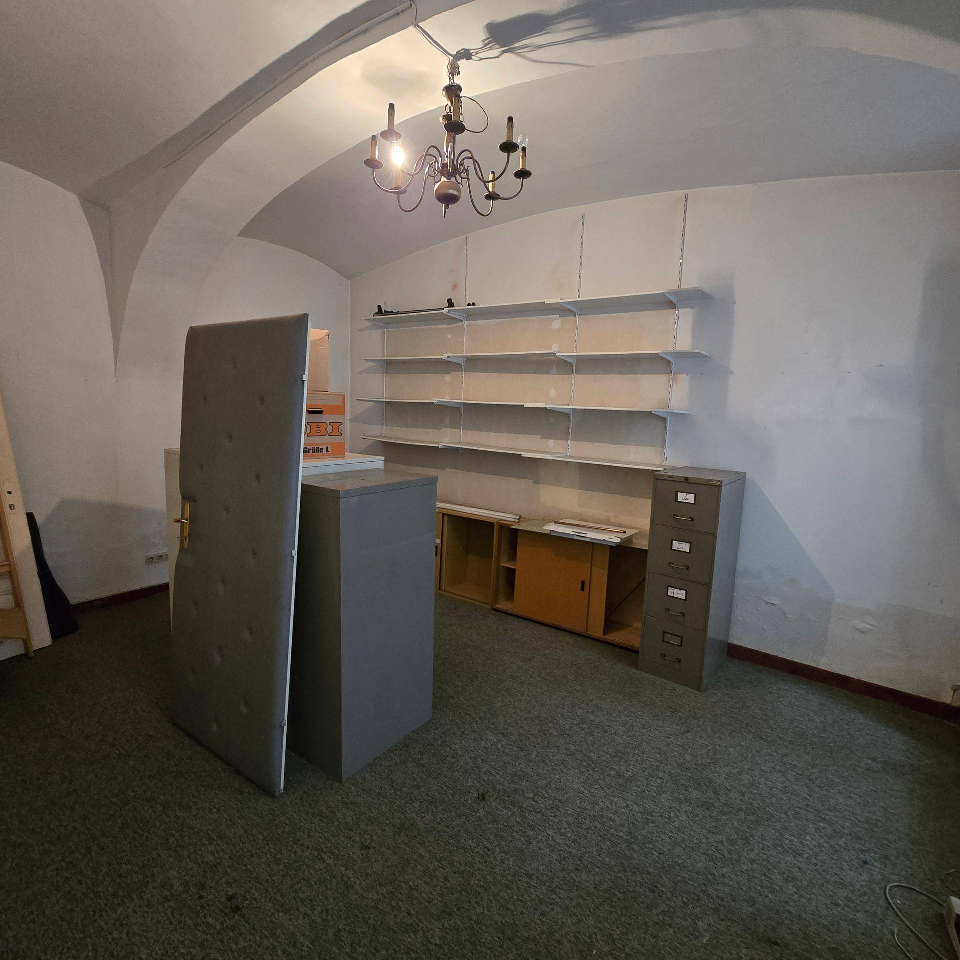 Büroraum mit Teppichboden, gewölbter Decke und Regalsystemen an der Wand, beleuchtet von einem Kronleuchter.