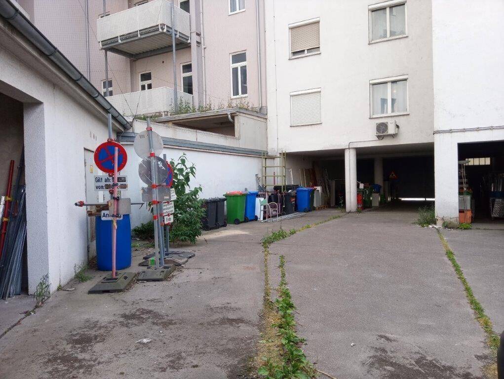 Außenansicht eines Gebäudes mit Zufahrt zu einer Tiefgarage und Mülltonnen am Rand.