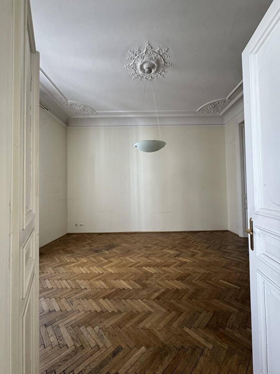 Leeres Zimmer mit Fischgrätparkett, hohen Decken und dekorativem Stuck an der Decke, bereit zur Einrichtung.