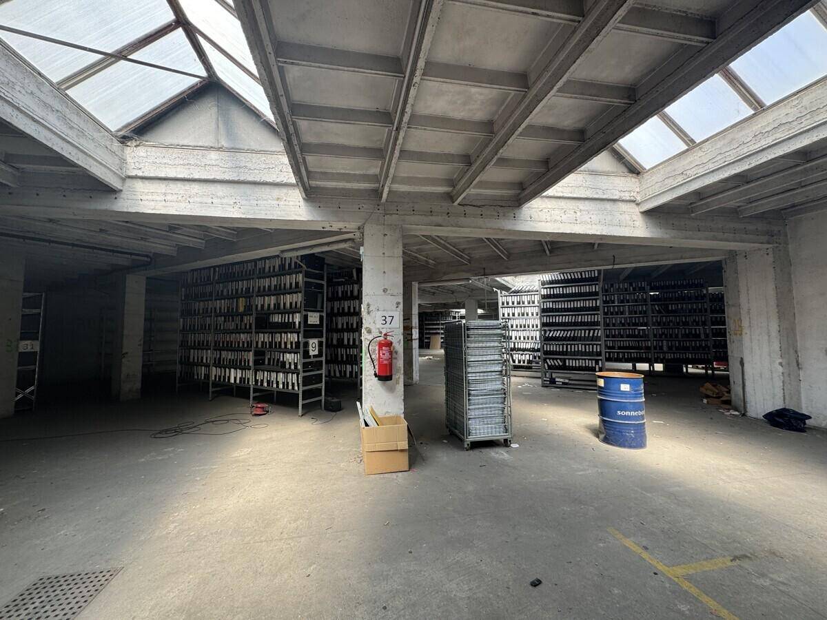 Lagerhalle mit Regalen voller Flaschen, Betonstützen und Oberlichtern, die Tageslicht hereinlassen.