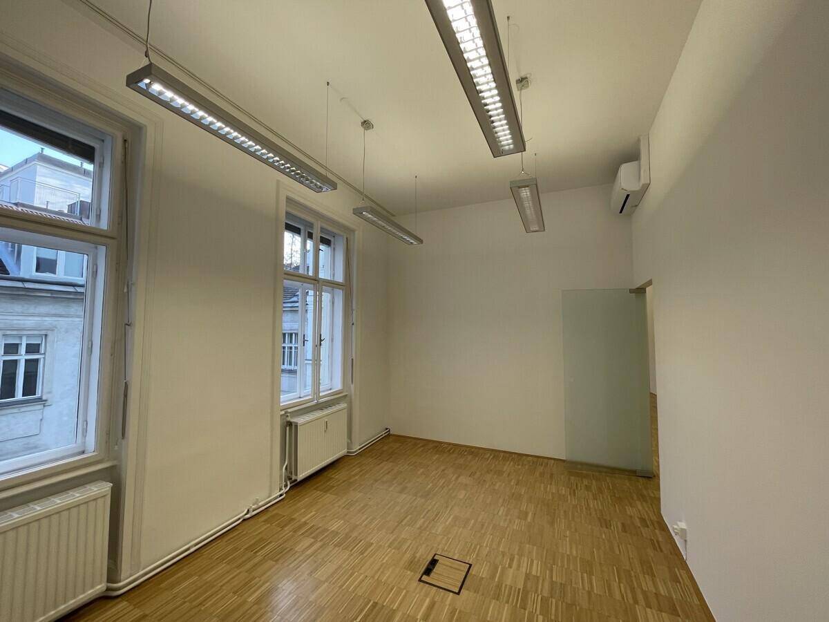 Heller Büroraum mit Parkettboden, großen Fenstern und moderner Deckenbeleuchtung.