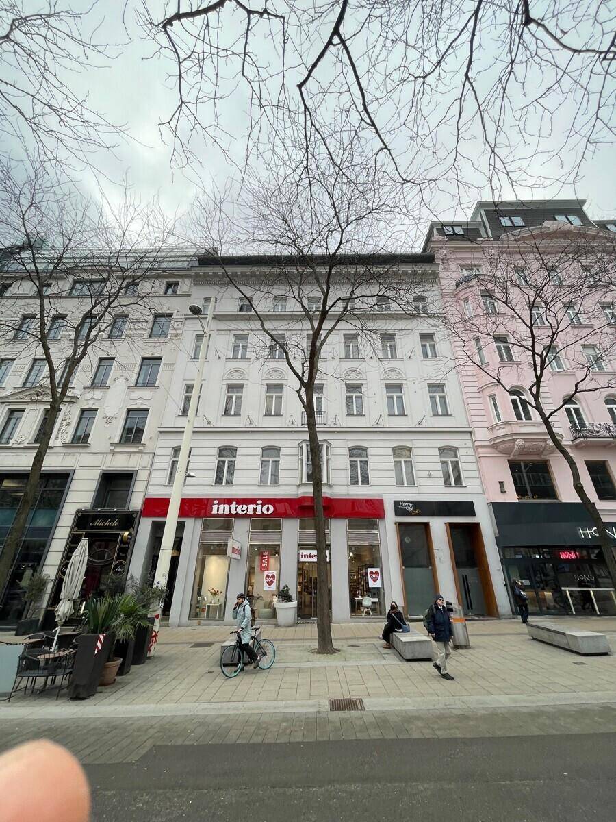 Gebäudeansicht von der Straße mit Geschäften im Erdgeschoss und Wohnungen darüber, umgeben von kahlen Bäumen.