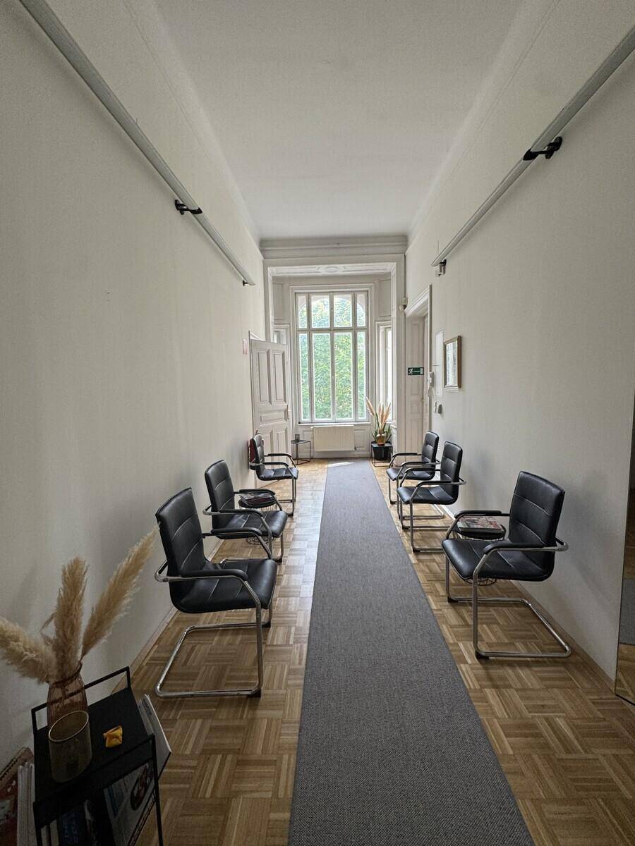 Langer Flur mit Fischgrätparkett, einer Reihe von Stühlen und einem großen Fenster am Ende, das Licht spendet.