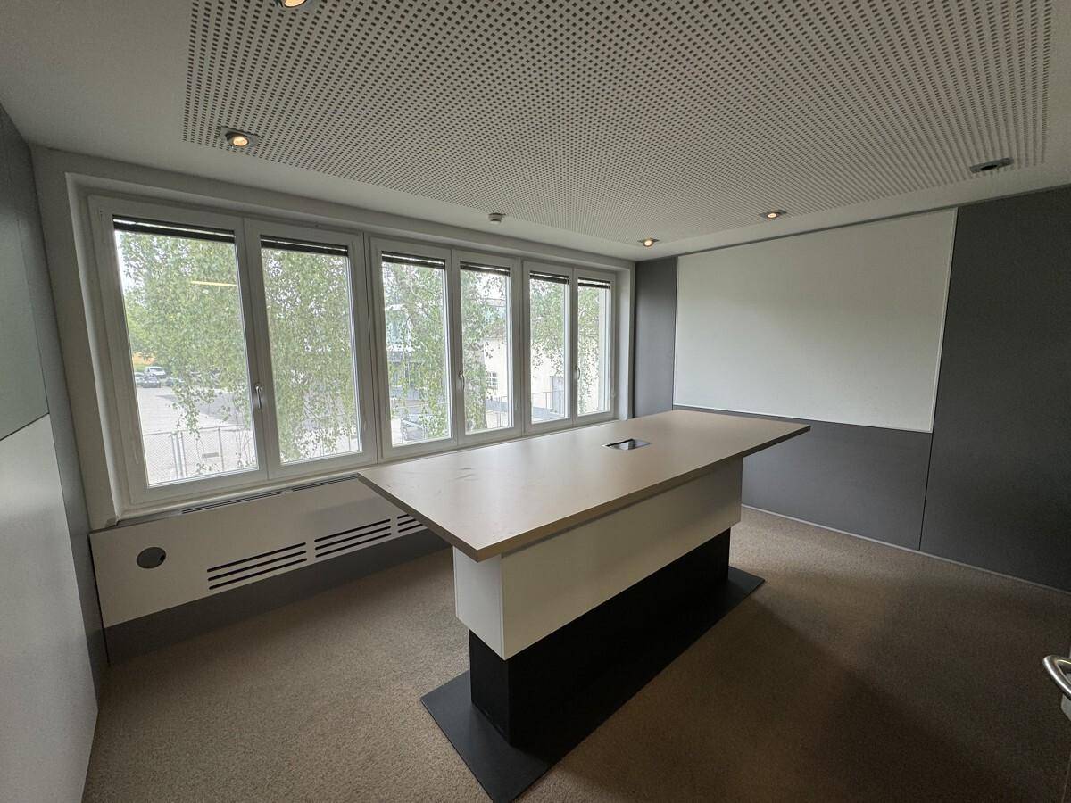Konferenzraum mit großem Tisch, Teppichboden und großen Fenstern, die viel Tageslicht hereinlassen.