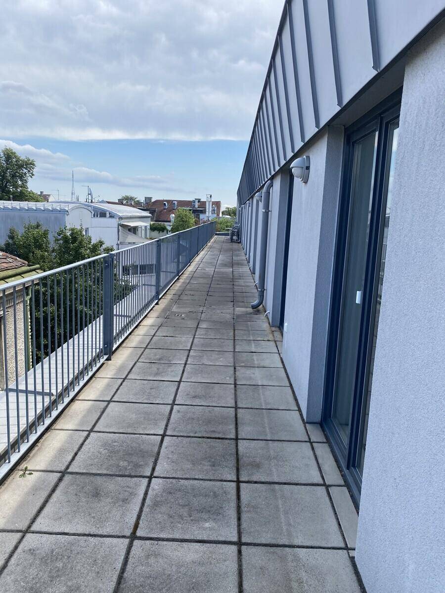 Lange, geflieste Dachterrasse mit Metallgeländer und Blick auf die umliegenden Gebäude und den Himmel.