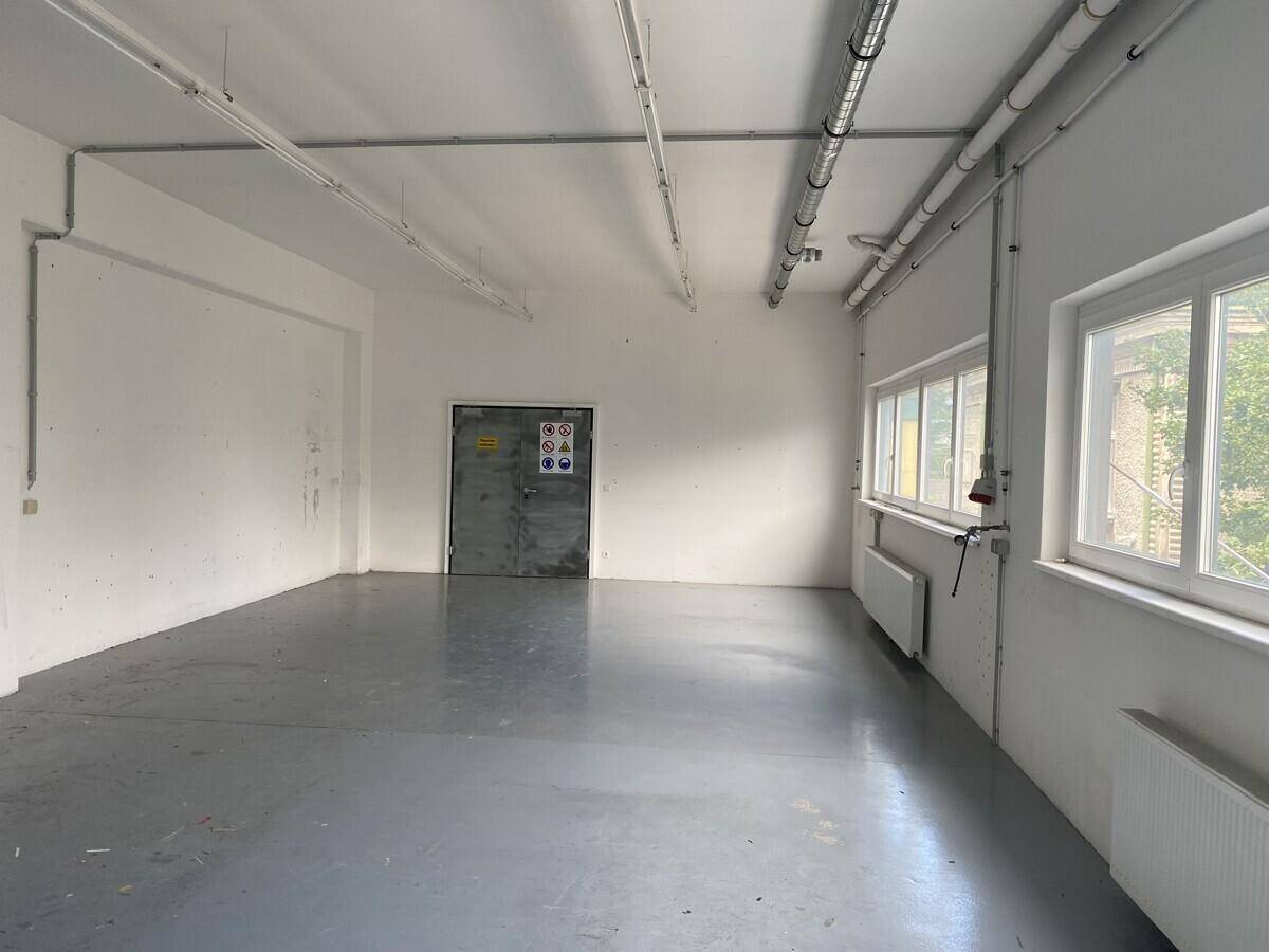 Leerer Industrieraum mit sichtbaren Rohren an der Decke, einem Metalltor und großen Fenstern, ideal für Lagerung oder Werkstatt.