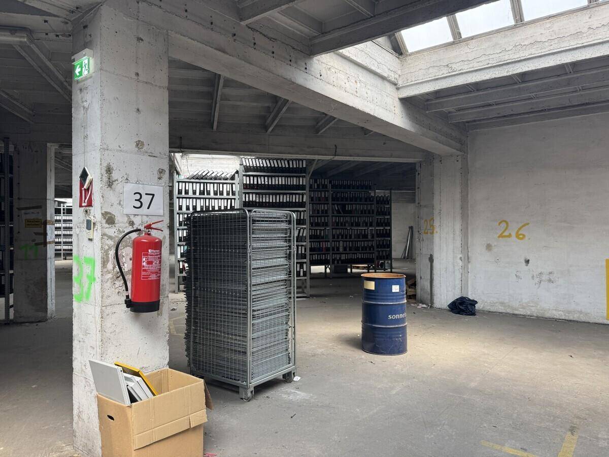 Großer, offener Lagerraum mit Betonstützen, Oberlichtern und Regalen, dazu ein Feuerlöscher und ein Fass.
