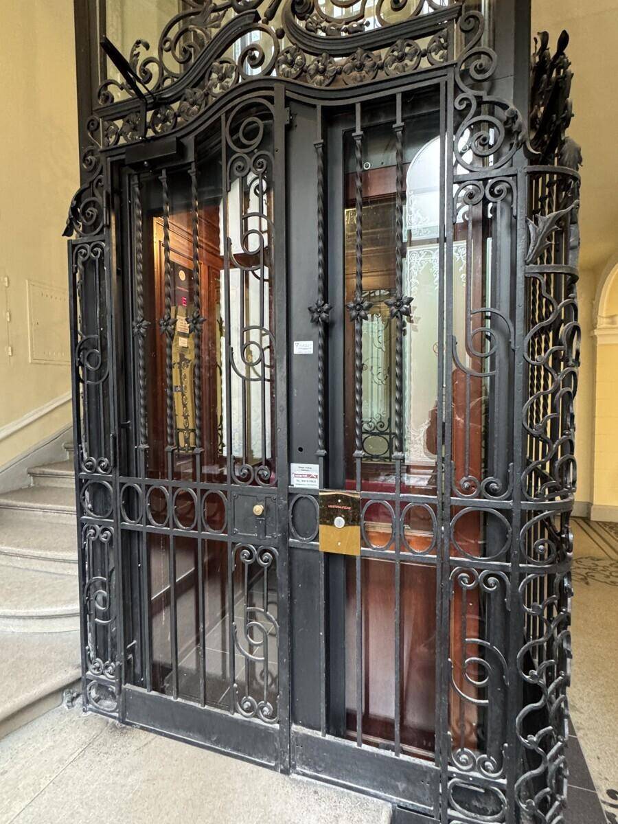 Historischer Aufzug mit kunstvollem schmiedeeisernem Gitter und Holzkabine in einem eleganten Treppenhaus.