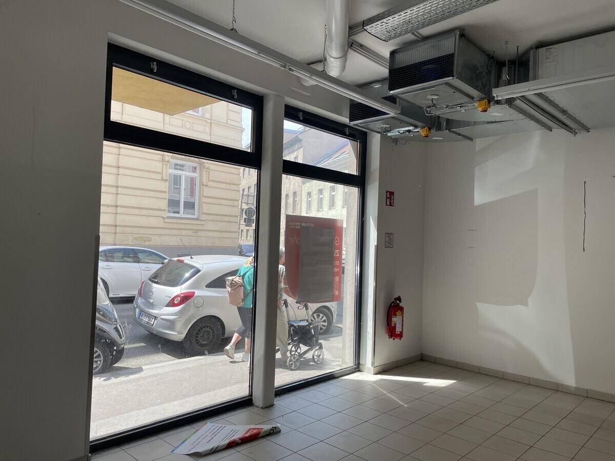 Kleinerer Gewerberaum mit großen Schaufenstern zur Straße, Fliesenboden und sichtbaren Installationen.