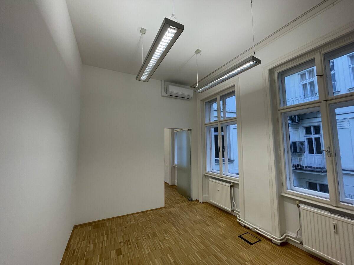 Büroraum mit Parkettboden, großen Fenstern und Zugang zu weiteren Bereichen.