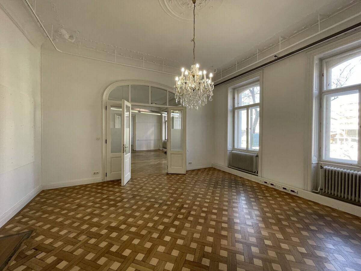 Eleganter Raum mit Fischgrätparkett, Kronleuchter und hohen Fenstern, die viel Licht hereinlassen.