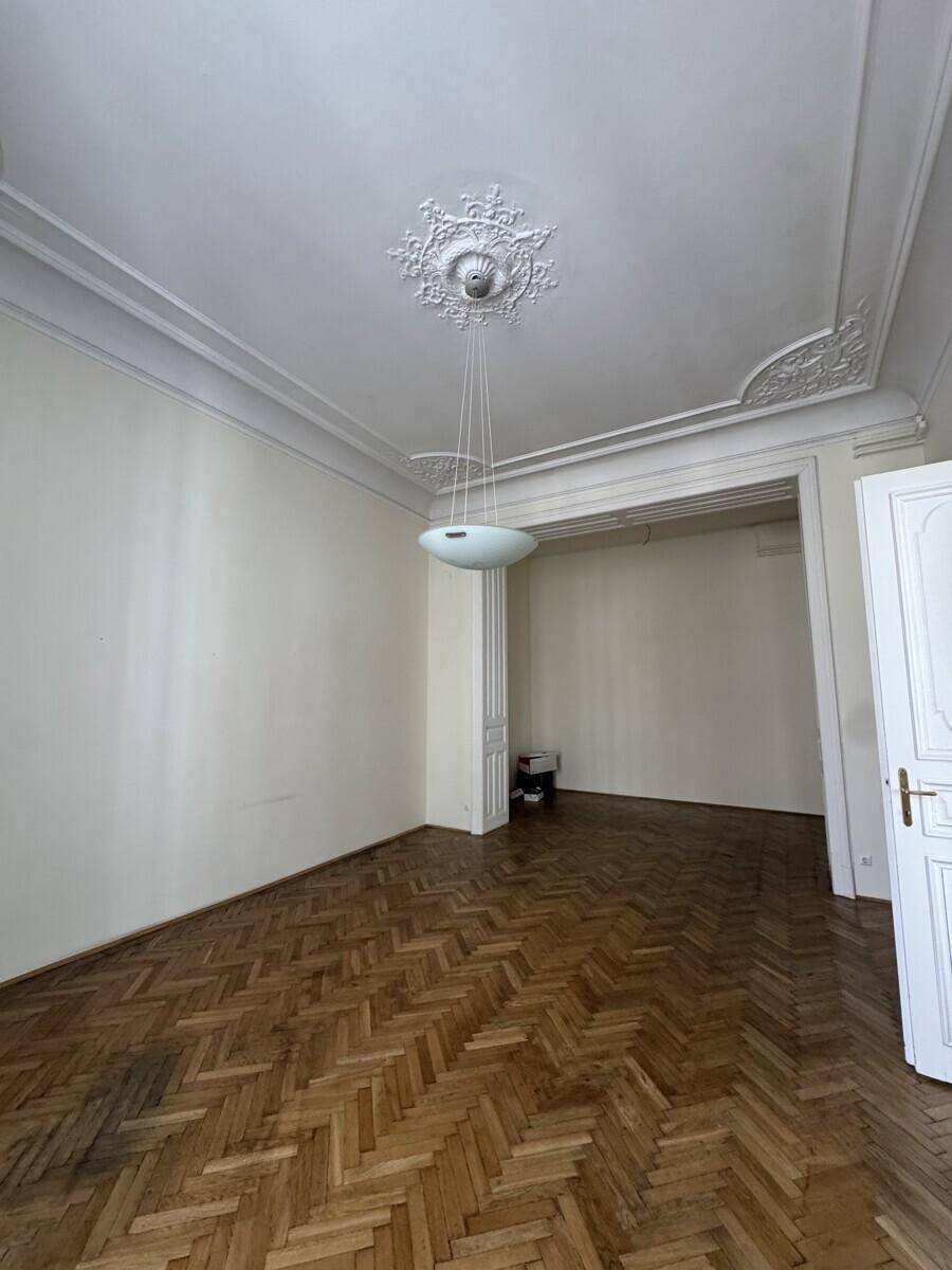 Helles Zimmer mit Fischgrätparkett, hohen Decken und einem dekorativen Stuckmedaillon an der Decke.