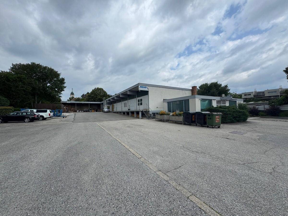 Industrielles Gebäude mit Laderampen und großem Parkplatz, umgeben von Bäumen und Büschen.