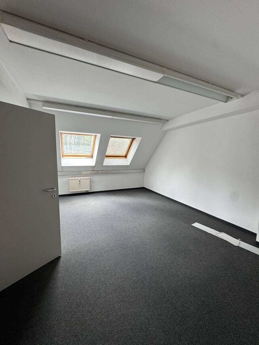 Leeres Büro im Dachgeschoss mit Teppichboden, schrägen Wänden und zwei Dachfenstern für natürliches Licht.
