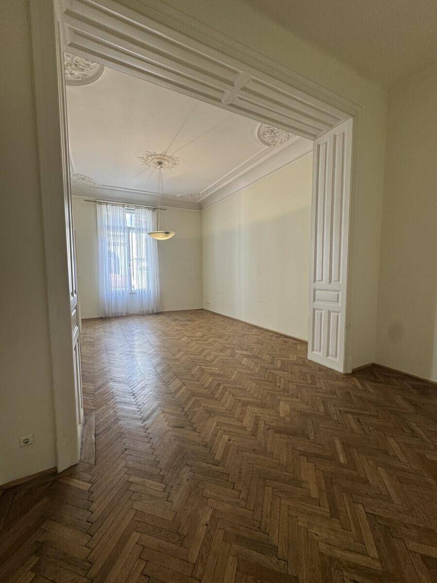 Geräumiges Zimmer mit Fischgrätparkett, hohen Decken und einem großen Fenster mit transparenten Vorhängen.