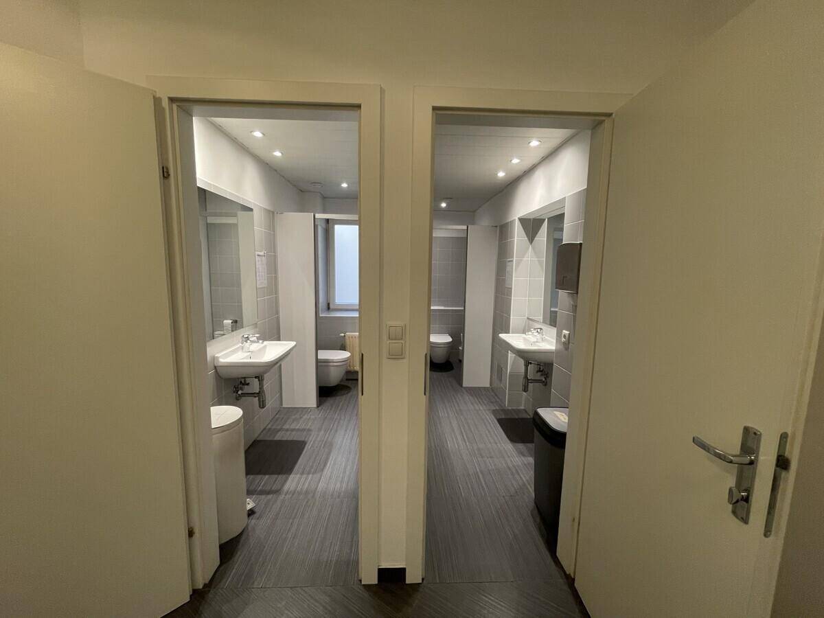 Zwei moderne Badezimmer mit Waschbecken, Toiletten und hellen Fliesen, durch offene Türen sichtbar.