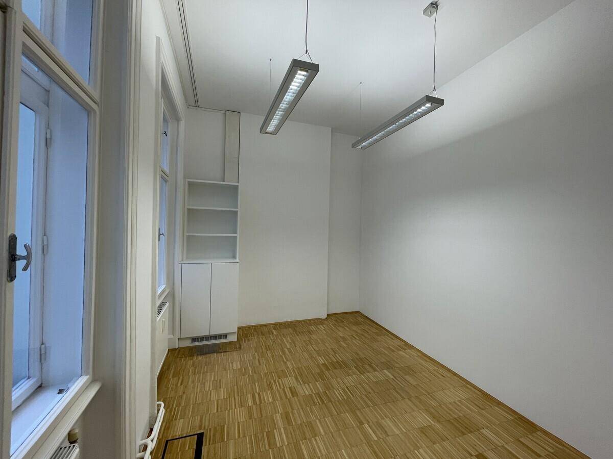 Kleinerer Büroraum mit Parkettboden, Einbauschrank und großen Fenstern.