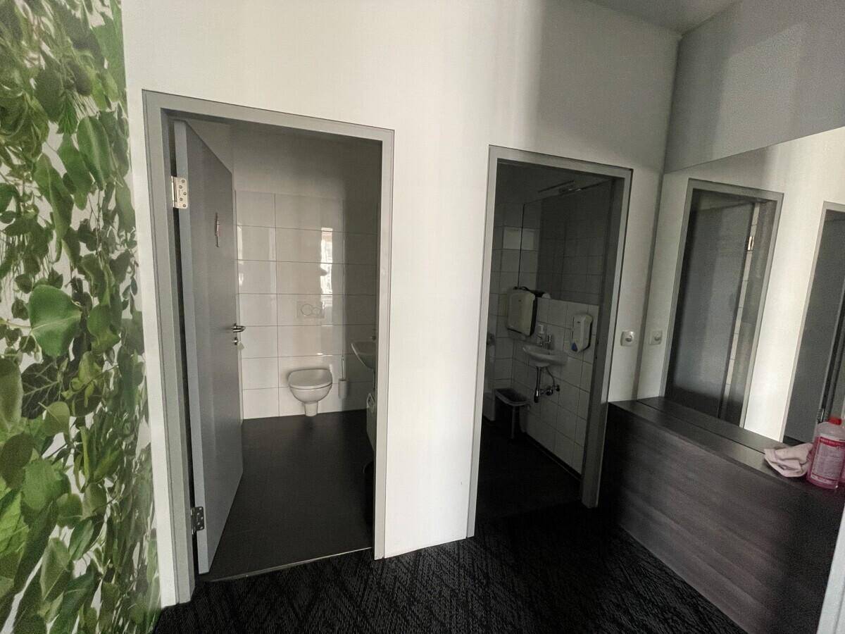 Zwei separate Toilettenräume mit weißen Fliesen, Waschbecken und dunklem Bodenbelag.