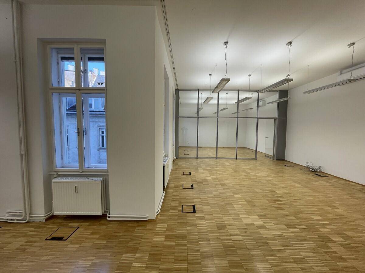 Großer Büroraum mit Parkettboden, hohen Fenstern und modernen Glasabtrennungen.