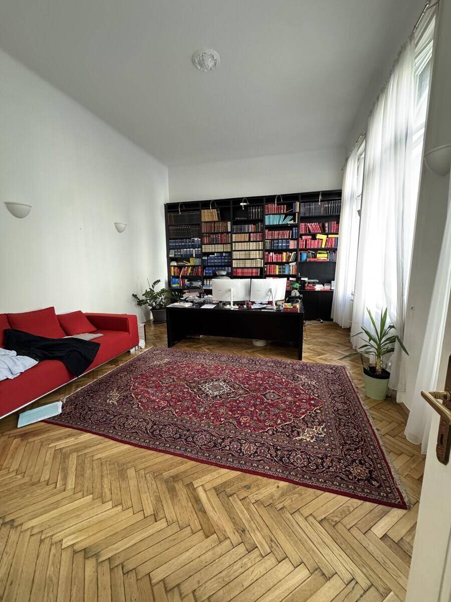 Geräumiges Büro mit Fischgrätparkett, großen Bücherregalen, einem Schreibtisch und einem roten Sofa.