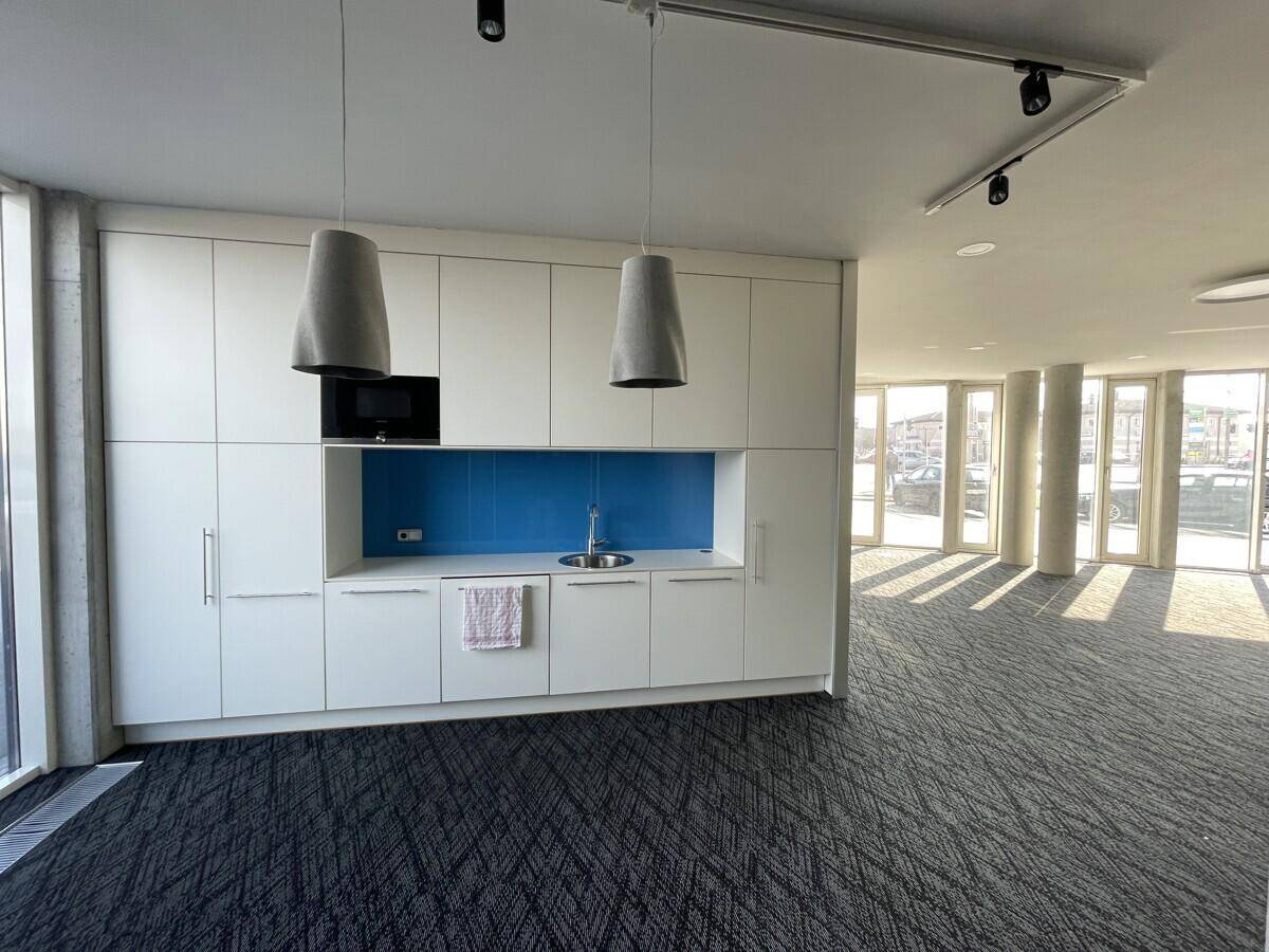 Moderne Teeküche mit weißen Schränken, blauer Rückwand und integrierten Geräten in einem offenen Bürobereich.