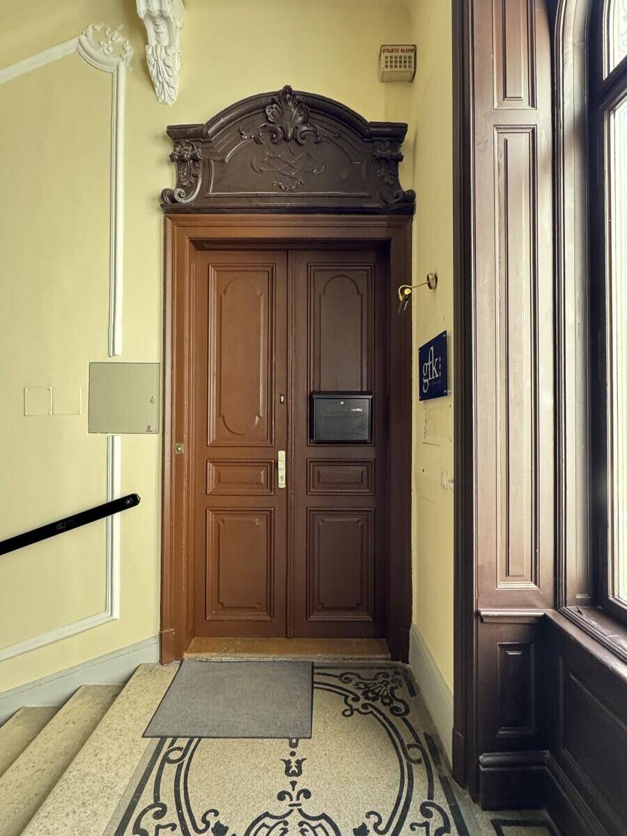 Prunkvolle braune Eingangstür mit Holzschnitzereien und gemustertem Fliesenboden im historischen Treppenhaus.