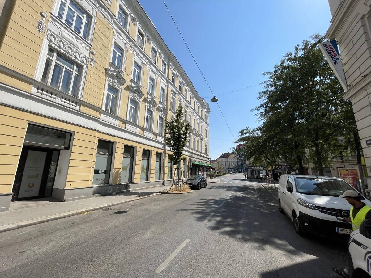 Eine belebte Stadtstraße mit historischen Gebäuden, Bäumen und geparkten Fahrzeugen unter blauem Himmel.